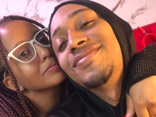 LilithAndAsmodeo with glasses, braided hair, and brown skin leans on a black-hoodie-wearing, bearded Asmodeo, smiling. Asmodeo's arm is around LilithAndAsmodeo's shoulder.