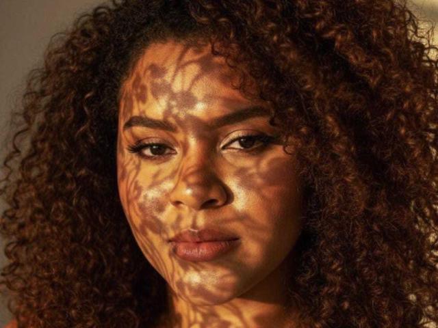 AkiraHeat has curly brown hair, her face is covered in shadow patterns, and she's staring with a serious expression, showcasing her full lips and dark eyes.