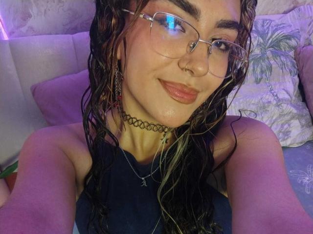 AdaraAngel con el cabello trenzado y mojado, gafas, piercing en la nariz, collar negro, y lápiz labial, sonriendo, mostrando sus pechos, vistiendo una blusa negra, sentada en una cama con luces moradas..