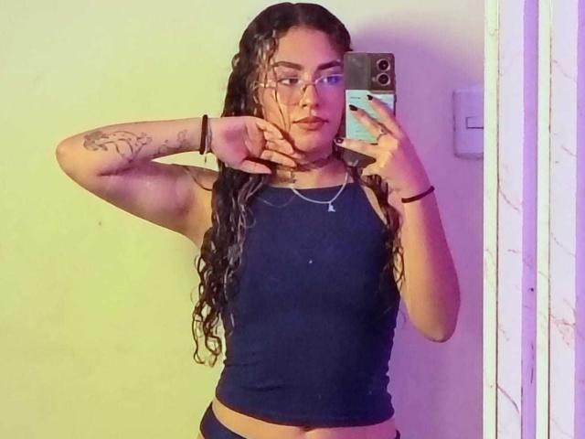 AdaraAngel, con el pelo largo, mojado y trenzado, vistiendo una camiseta negra, mostrando un tatuaje en su brazo derecho, tomando una foto de sí misma en el espejo, mostrando sus pechos pequeños y planos..