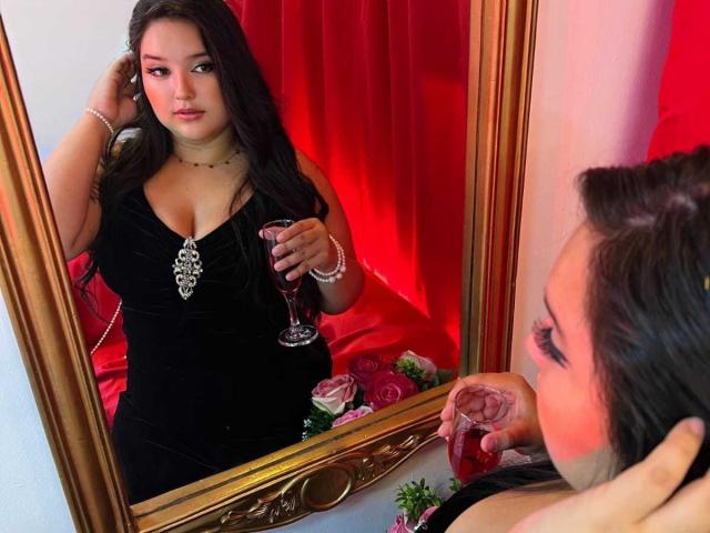 RositaBennet with long dark hair, wearing a black dress with a diamond necklace, holding a red wine glass, reflected in a gold-framed mirror, showing her tits and face.
