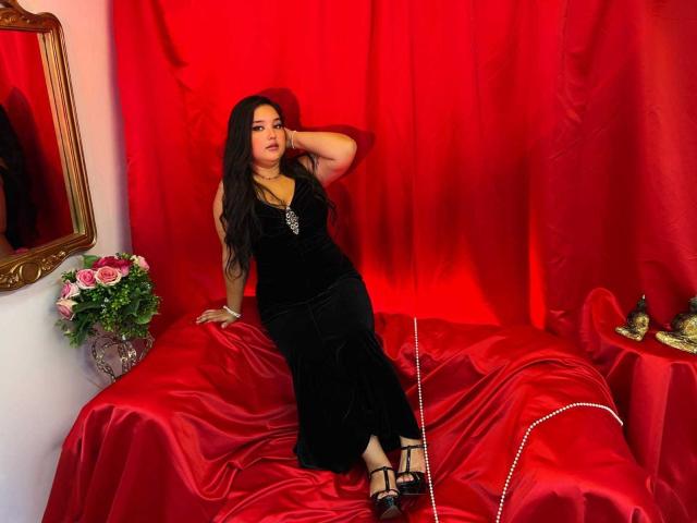 RositaBennet with long black hair, wearing a black velvet dress, sitting on red satin, her hand on her head, black heels, red curtains, flower vase, mirror, and jewelry.
