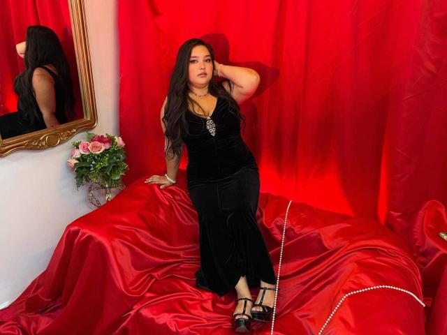 RositaBennet with long dark hair, black velvet dress, and black heels, sitting on red satin, hand in hair, mirror reflection, bouquet of pink roses on left.