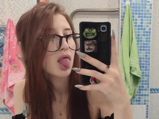AuroreProvosty with long red hair, black glasses, and long gray nails, sticking her tongue out, taking a mirror selfie with a black phone, in a bathroom.