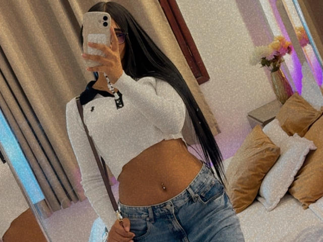 AntonellaPreschot with long black hair, wearing a white crop top, showing her tits, and blue jeans, holding a phone, with a navel piercing, standing in a room.