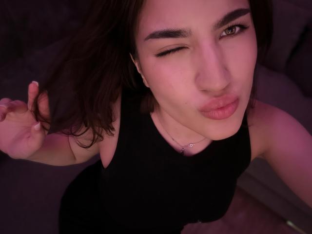 EstherFleitman with one eye closed, puckering lips, dark brown hair, black sleeveless top, necklace, holding hair, pinkish-purple lighting, focused on face, slightly upward angle, casual selfie.