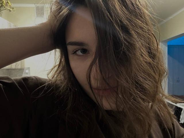 EstherFleitman with messy brown hair covering one eye, hand in hair, subtle smile, wearing a dark shirt, looking at camera, blue door in background.