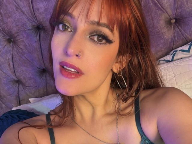 JaneRoa with red hair, green eyes, black eyeliner, pink lipstick, nose piercing, hoop earrings, green bra, necklace, lying on bed, looking at camera, slightly open mouth, purple lighting.