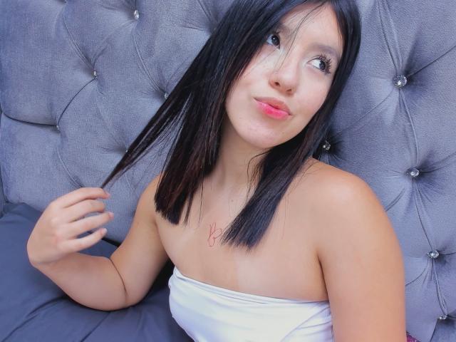 AdelleVelvet, con cabello negro lacio, piel clara, labios rosados, senos pequeños, vestida con un top blanco sin tirantes, sujetándose el cabello, sentada en una cama gris con cabecera tapizada..