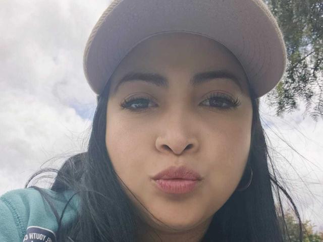 AlanaWest has dark eyes, long black eyelashes, and pink pouty lips. She's wearing a pink cap and green hoodie, with a nose piercing. Her black hair is loose.