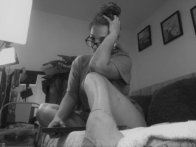 AmberDemons, sitting on couch, wearing glasses, curly hair in bun, touching head, in t-shirt, socks, anklet, crossed legs, holding phone, monochrome, slightly messy, cozy room.