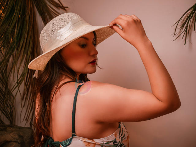 LunaPleasure with her hand on a white straw hat, showing her shoulder tattoo and green strap, her tits slightly visible, brown hair, red lipstick, and a cute face.