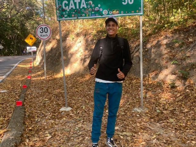 JackFox69, standing with thumbs up, wearing black hoodie, blue jeans, white tee, and black cap, on leaf-covered road, next to 30 km/h speed sign, and green road sign.