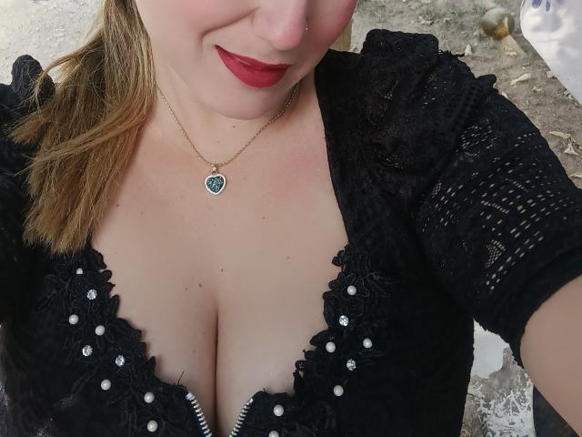 AlexaHotSeins com seios grandes, vestindo um vestido preto de renda, com botões de pérola, batom vermelho e um colar de coração verde, mostrando o decote. Ela tem pele clara e um piercing no nariz..