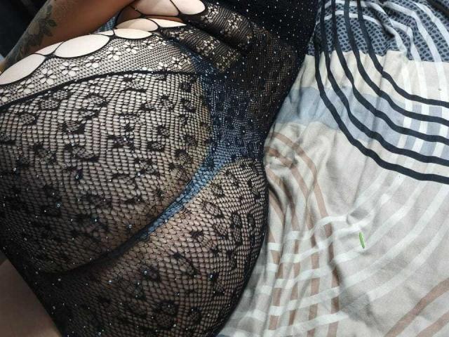 AlexaHotSeins indossa un abito a rete nero e scintillante con inserti tagliati, che mette in mostra il suo grande sedere e un tatuaggio sul braccio superiore. Sullo sfondo, una lenzuola a righe..