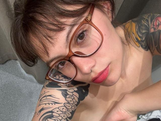 IsabellaArias with brown glasses, short brown hair, pink lips, tattoos on her arm, looking up, topless, showing her small tits, holding her chest.
