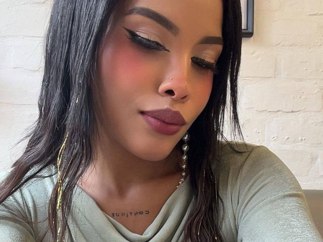 LexieBloom has dark brown hair, winged eyeliner, pink blush, purple lipstick, and a nose ring. She's wearing a green top with gold earrings. Her tits are not visible.