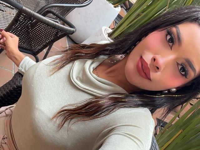 LexieBloom with her tits in a tight, white, long-sleeve top, pink blush, red lipstick, and brown wavy hair, holding a chair, looking at the camera.