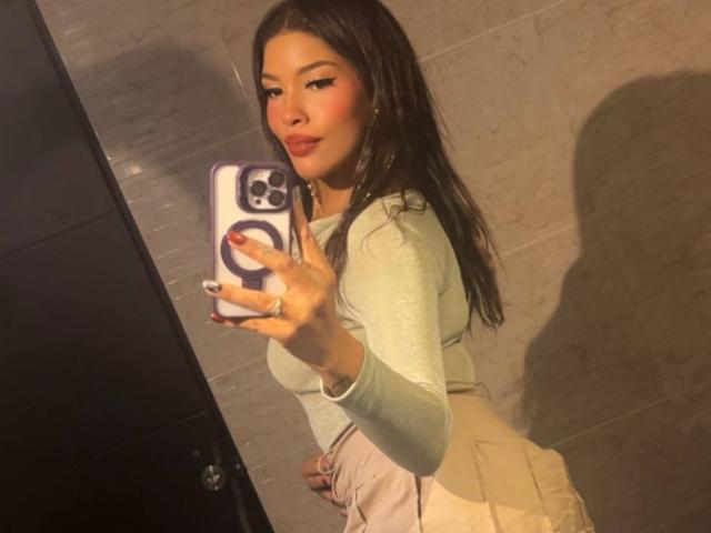LexieBloom with long black hair, green top, and beige pants, taking a mirror selfie with a phone, showing her ass and a sexy smile.