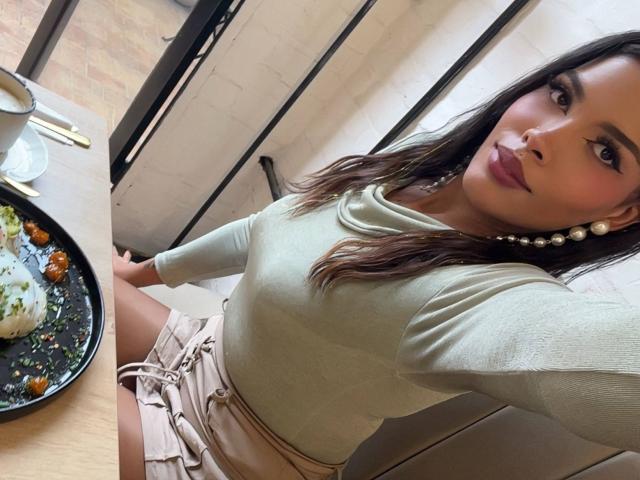 LexieBloom with brown hair, wearing a green top, pearl earrings, and beige shorts, sitting at a table with a plate of food. Her tits are small, and she's smiling.