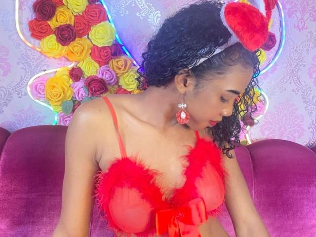 PerleNoireX with curly hair, red feathered bra, red heart headband, and red earrings, sitting on a purple couch, looking down, colorful floral wings in background.