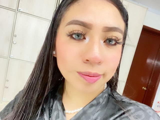 ZoeRedds hat langes, schwarzes Haar, grüne Augen, dicke Augenbrauen, rosafarbene Lippen und eine Perlenkette. Sie trägt ein schwarzes Hemd, unter dem ihre Brüste sichtbar sind. Ihr Make-up umfasst lange Wimpern..