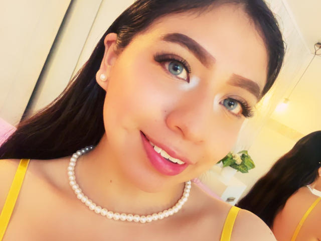 ZoeRedds has blue eyes, dark brown hair, and pink lipstick, wearing a pearl necklace and yellow dress, with a cute smile showing her white teeth.