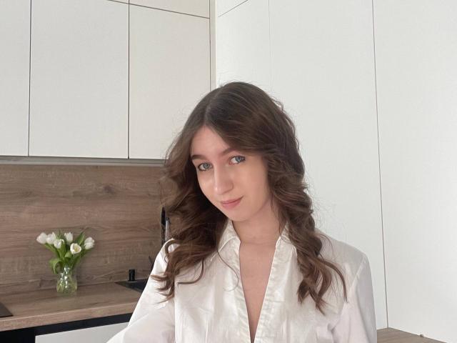 Sara Rosenbarger, con i capelli castano ondulati, gli occhi verdi e una camicia bianca con bottoni, sorride dolcemente in una cucina moderna. Armadietti bianchi e schienale in legno. Un piccolo vaso con tulipani bianchi..