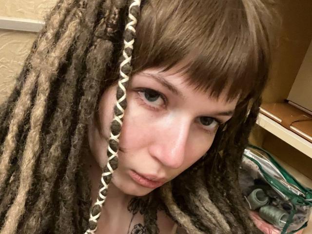 FerminaHilmer with brown dreadlocks, blue eyes, and a pensive expression, has a small nose and full lips. Her hair has a white beaded section.