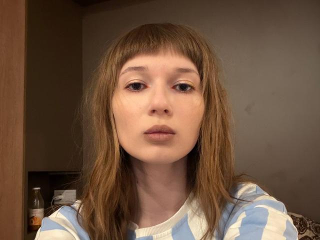 FerminaHilmer with straight brown hair, blue and white striped shirt, pale skin, and pouty lips, looking directly at camera. Soft lighting on her face.