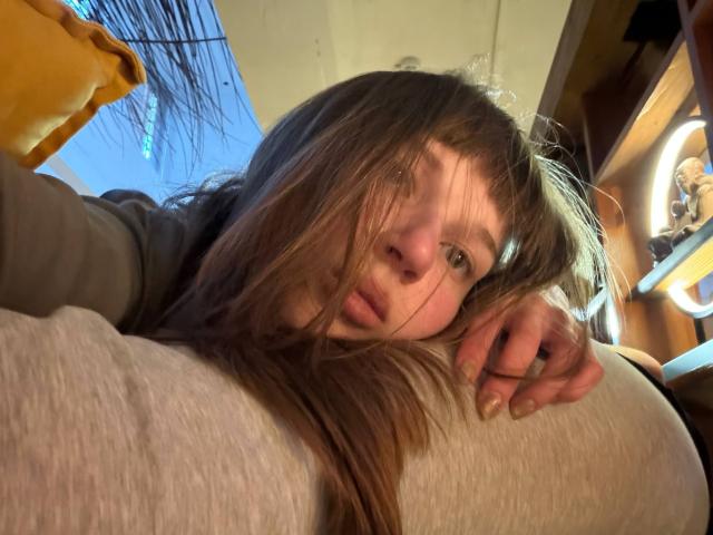 FerminaHilmer with messy brown hair, green eyes, and a tired expression, leaning on a gray pillow, wearing a green hoodie, right hand resting near her face.