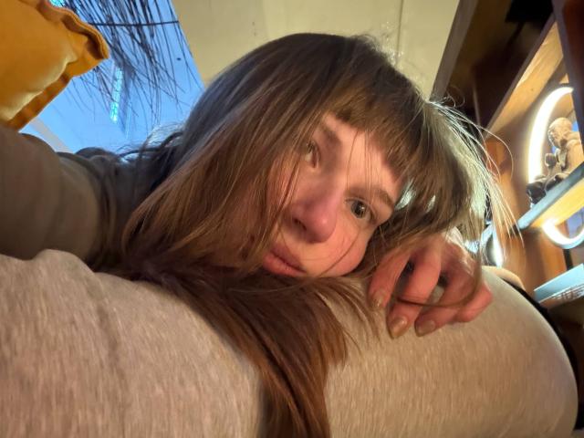 FerminaHilmer with messy brown hair, nose slightly red, lying on a pillow, eyes looking at camera, hand resting on pillow, wearing a gray shirt. Low-angle shot.