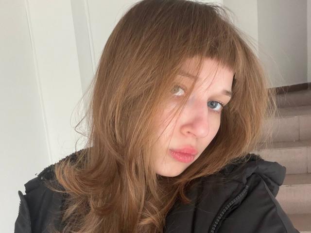 FerminaHilmer with messy brown hair, blue eyes, and pink lips, wearing a black jacket, looking slightly to the side with a subtle pout. Soft natural light.