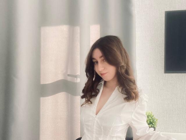 Sara Rosenbarger tiene el pelo castaño ondulado, una camisa blanca con botones y una sonrisa encantadora, y está de pie cerca de una cortina con un televisor de pantalla plana a la derecha..