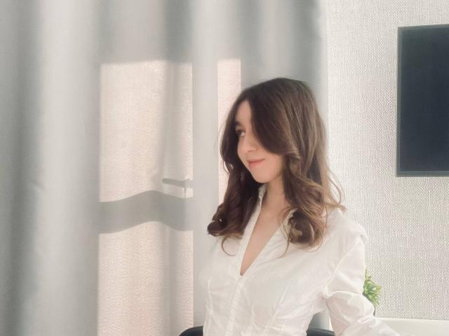 Sara Rosenbarger, avec ses cheveux bruns ondulés, portant une chemise blanche à boutons, regardant sur le côté avec un sourire discret, se tenant devant un rideau..
