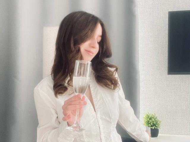 Sara Rosenbarger, avec des cheveux bruns ondulés, tenant une flûte de champagne, portant une chemise blanche à boutons, avec un léger sourire, éclairage doux, petite plante verte sur la droite..