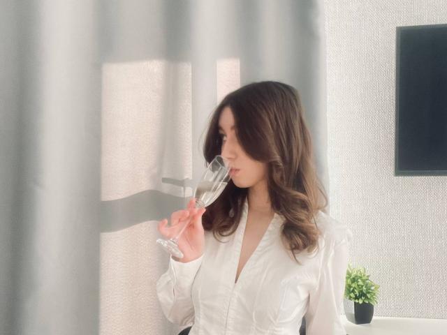 SaraRosenbarger with wavy brown hair, white shirt, sipping from a clear glass, standing by a curtain, holding it to her lips, looking to the side.