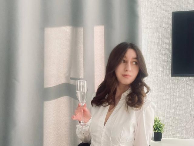 Sara Rosenbarger mit welligem, braunem Haar, einem weißen Hemd mit Knopfleiste, die ein Champagnerglas hält und in der Nähe eines Vorhangs steht. Auf einem Tisch steht eine kleine, topfgebundene Pflanze. Sie lächelt leicht..