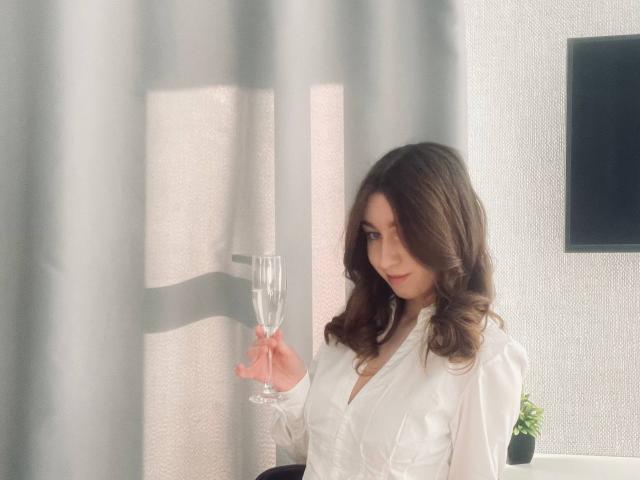 Sara Rosenbarger, met golvend bruin haar, een witte satijnen badjas, een champagneglas in haar hand, een subtiele glimlach, staat voor dunne grijze gordijnen en kijkt iets opzij..