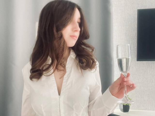 Sara Rosenbarger mit welligem, braunem Haar, einem weißen Hemd mit Knopfleiste, die ein Champagnerglas hält, lächelt, schaut zur Seite und steht neben einer kleinen, grünen Pflanze..