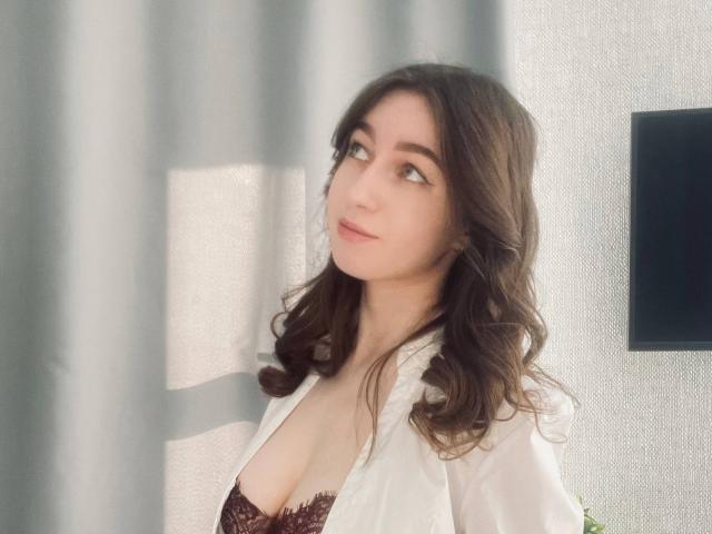 Sara Rosenbarger, met golvend bruin haar, draagt een witte blouse en een bordeauxrode kanten beha, kijkt opzij met een lichte glimlach en laat haar borsten zien..
