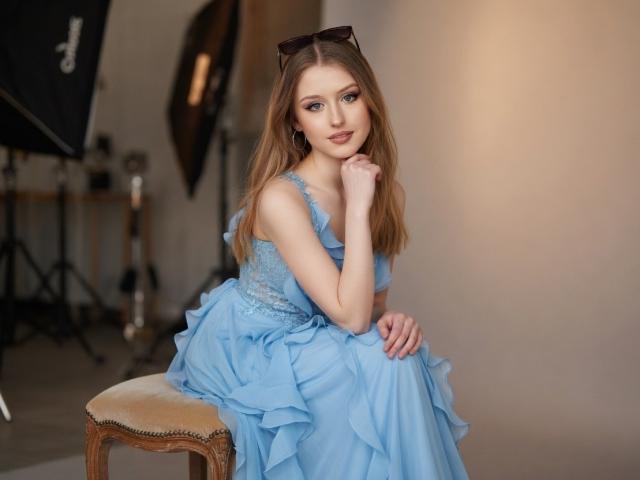 Nikollia, con un vestido azul, sentada en un taburete acolchado, con la mano cerca de la boca, cabello rubio largo, ojos azules, gafas en la cabeza, sonrisa suave, piel clara, expresión gentil..