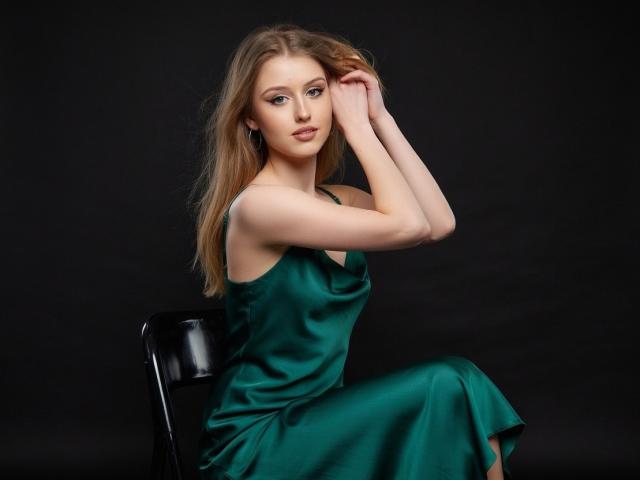 Nikollia, with long blonde hair, sits on a black chair, wearing a green satin dress, her arms raised, touching her hair, showing her tits, against a dark background.