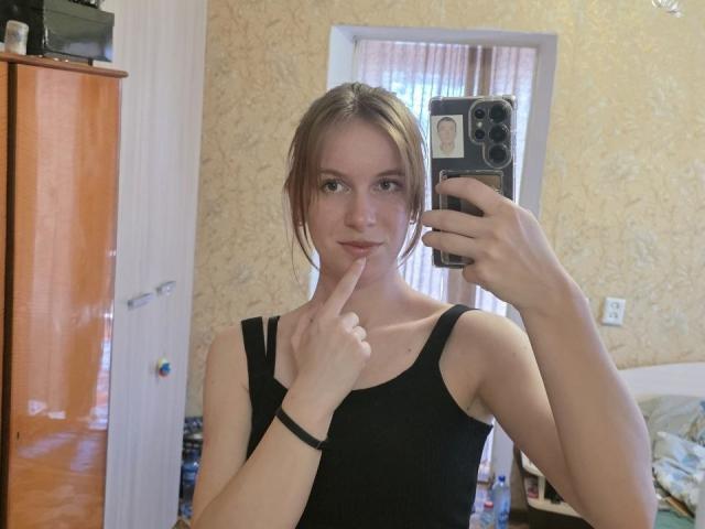 DebbyPennimpede with light brown hair, wearing a black tank top, holding a phone, finger on her bottom lip, mirror selfie, showing her small tits and slim build.