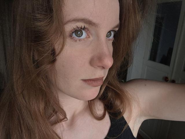 Debby Pennimpede, con ojos verde claro, cabello castaño ondulado y una expresión seria, lleva una blusa negra, mostrando su brazo y nariz desnudos. Tiene un rostro pequeño y con pecas..