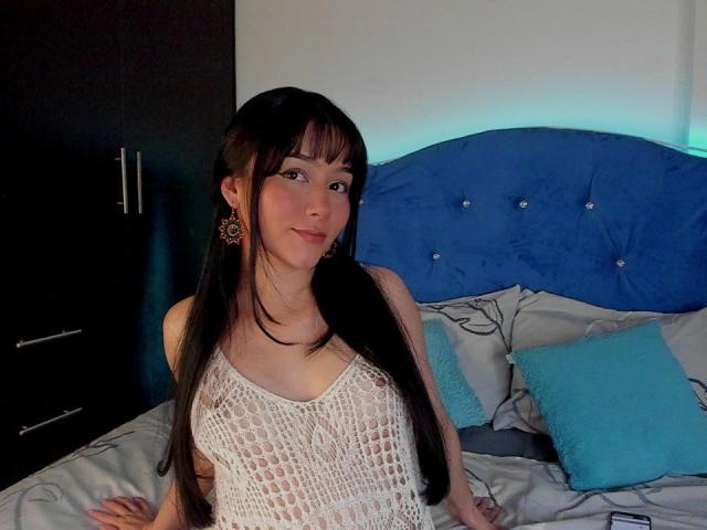 DeisyWest with long black hair, wearing a white, see-through knit top, showing small tits, blue earrings, sitting on a bed with a blue headboard and turquoise pillows.