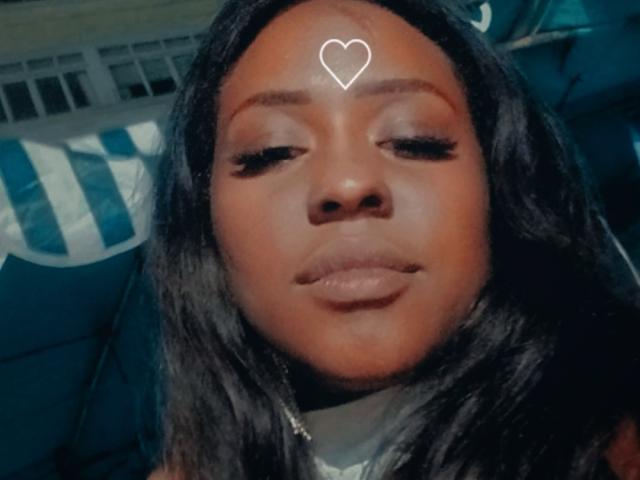 Thamires69, con piel morena, cabello negro largo y un adhesivo digital blanco en forma de corazón en la frente, tiene los ojos cerrados y los labios llenos, y está sentada en una silla azul..