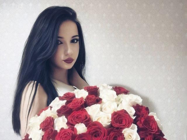 Gracia Ponting, con el pelo largo y negro, sosteniendo un ramo de rosas rojas y blancas, mirando por encima del hombro con ojos seductores y pintalabios rojo..