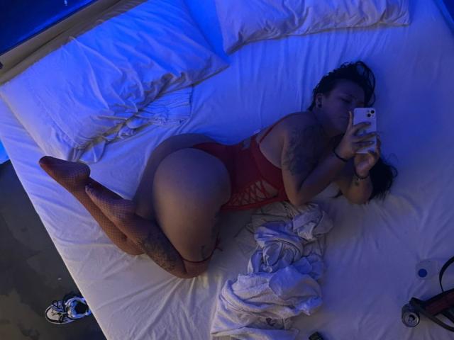 Una donna formosa, sdraiata su un letto, indossa una mutandina rossa con lacci, sta scattando un selfie con un telefono, mostrando tatuaggi, calze arancioni e il sedere, sotto una luce blu..