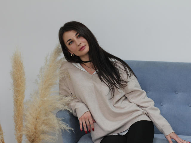 AlexOakman with long black hair, wearing a beige sweater and black pants, sits on a blue couch, puffy tan pampas grass to the left, red nails, slight smile.